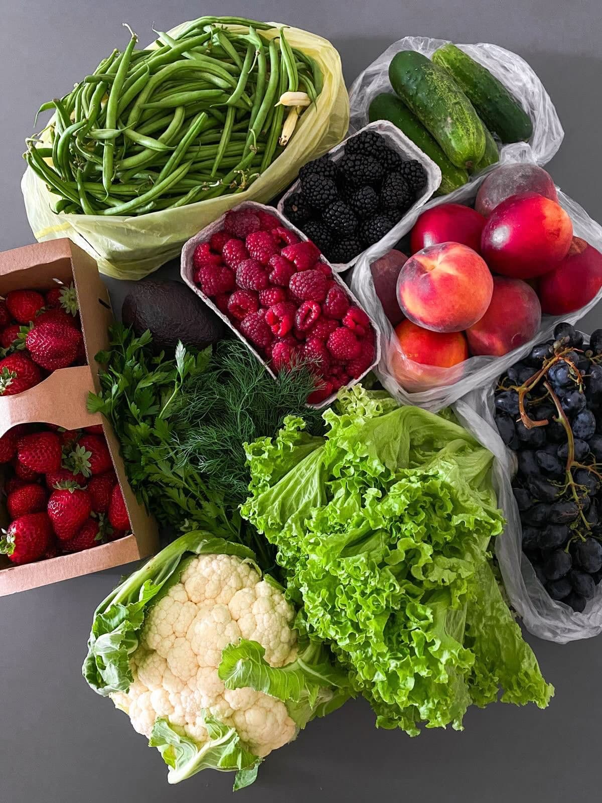 Assortimento di frutta e verdura fresca: fragole, pesche, lattuga, cavolfiore e fagiolini su sfondo grigio.
